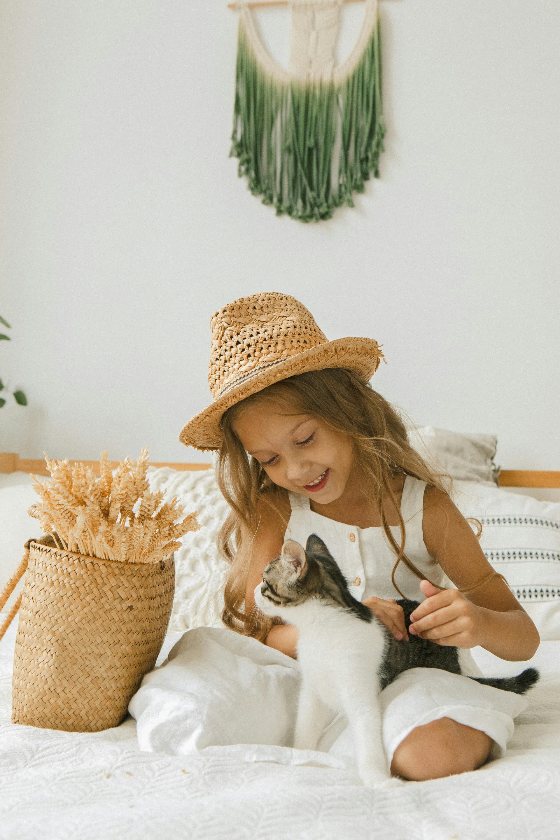 Child in a straw hat sitting on a bed and petting a kitten.